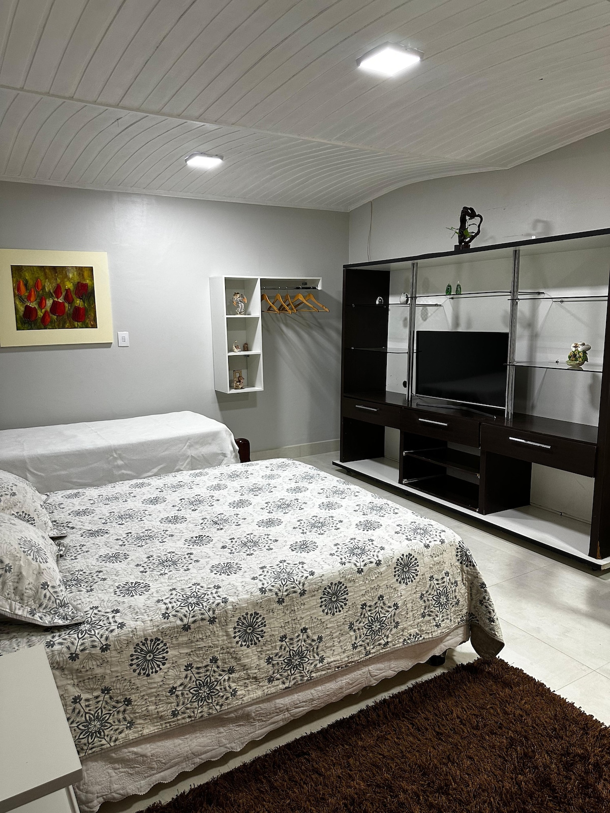 A well-lit sleeping area features a spacious bed covered in a patterned bedspread, complemented by a white sofa. A media unit with a television provides entertainment, while a small shelf displays decorative items. Wall-mounted storage holds hangers, enhancing organization.