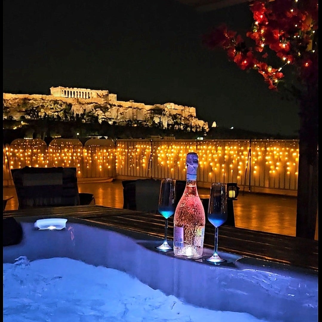 A jacuzzi is shown illuminated in soft blue light, accompanied by two glasses filled with drinks and a bottle of sparkling wine. The Acropolis is visible in the background, beautifully lit against the night sky, with twinkling lights adorning the terrace.