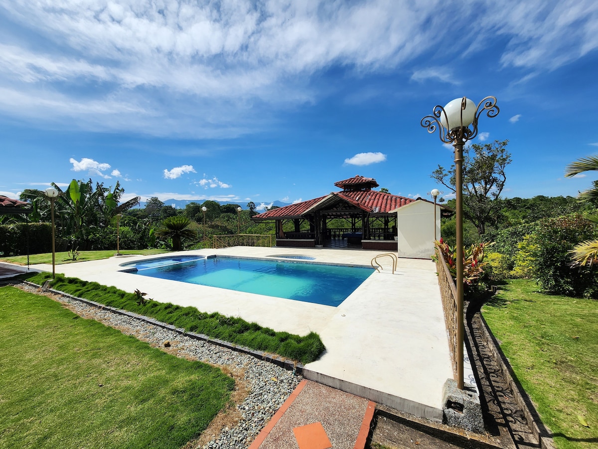 An outdoor area features a rectangular swimming pool surrounded by a well-manicured lawn. A covered seating area with a tiled roof is visible in the background, complemented by clear blue skies and a few scattered clouds.