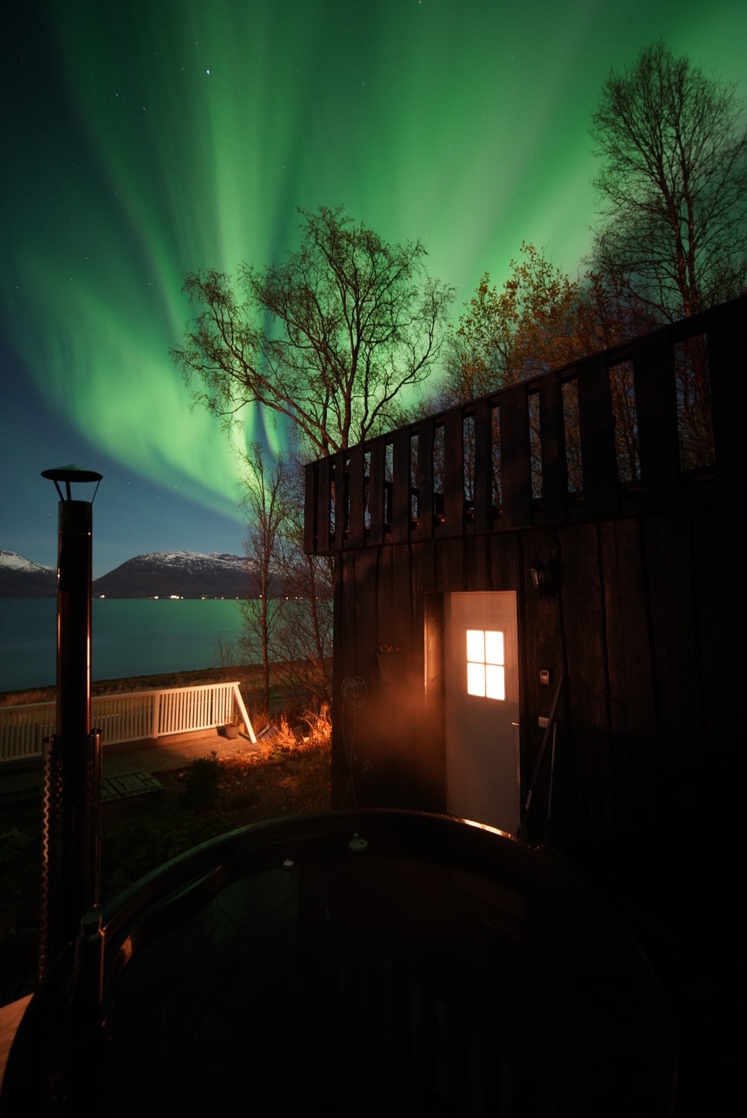 The image captures the vibrant Aurora Borealis lighting up the night sky above a dark wooden guesthouse. A soft glow emanates from a window, illuminating the entrance, while a hot tub is visible in the foreground. Scenic mountains and water are framed in the background.