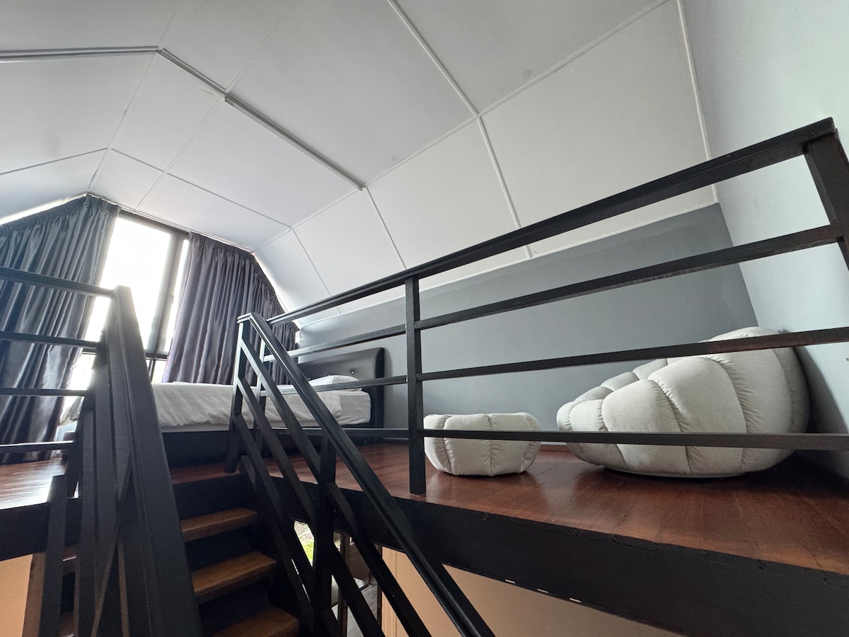 A spacious loft area features a cozy sleeping space with a bed positioned beneath a high ceiling. A comfortable seating area includes soft, cushioned chairs. Natural light filters through large windows, complemented by dark curtains, creating a serene and inviting environment.