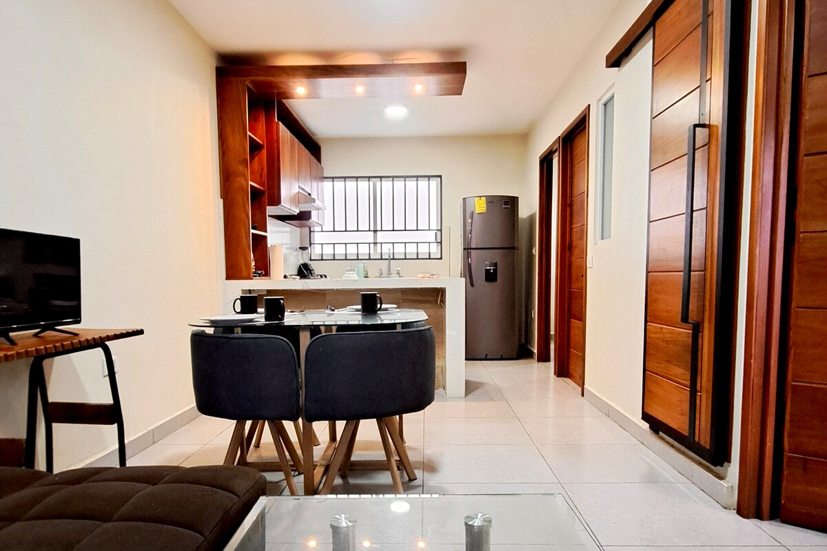 A modern living area showcases a dining table with four chairs, complemented by a sleek countertop. The kitchen area features quality cabinetry, and a refrigerator is visible in the background. Light-colored tiles reflect natural light, enhancing the spacious layout.