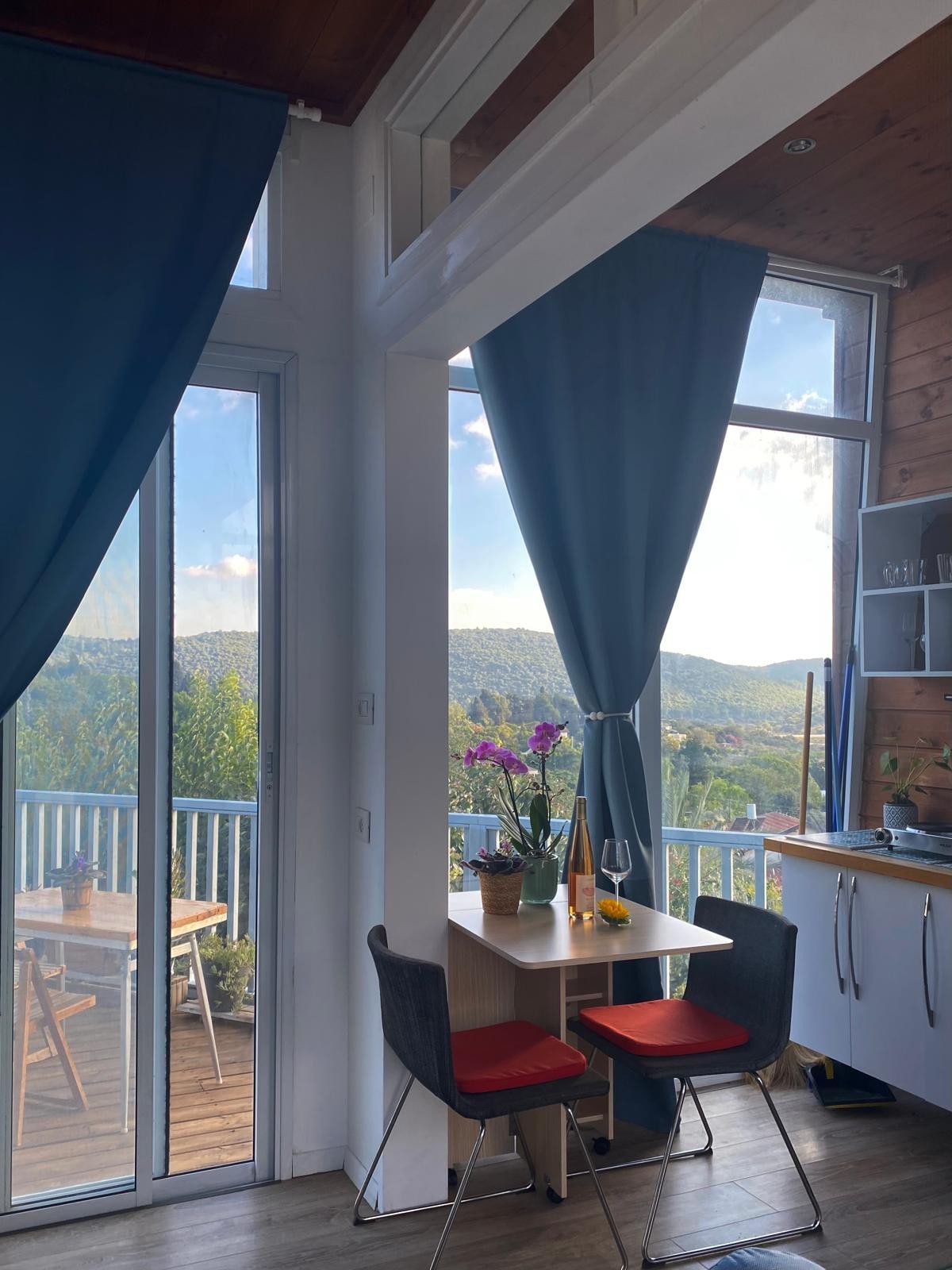 A dining area is visible with a small table and two chairs, accented by red cushions. A potted orchid sits at the center of the table. Large windows are framed by blue curtains, revealing a scenic view of surrounding hills.