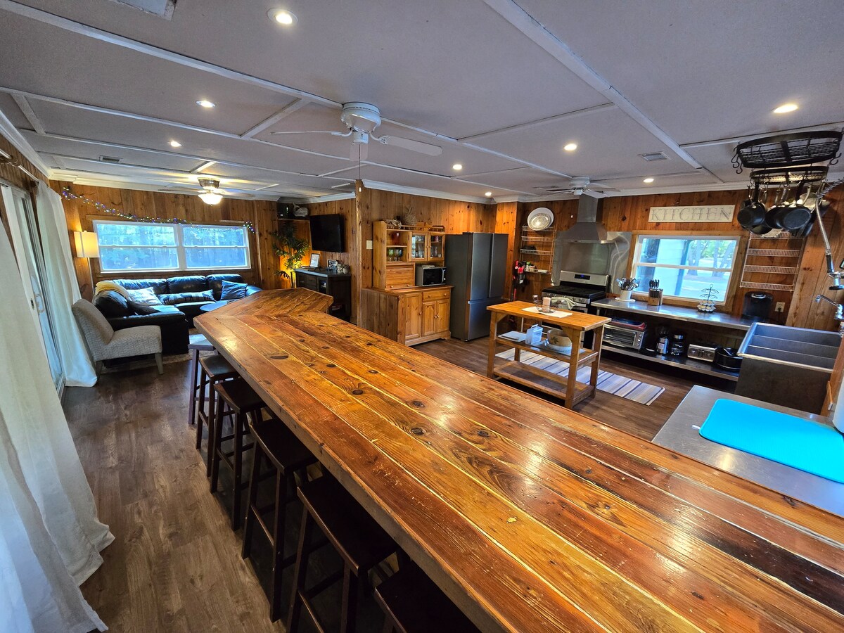 The spacious kitchen features a large wooden bar with seating for seven. A fully equipped cooking area is visible, including modern appliances, a dining table, and ample counter space. Natural light fills the room through windows, complementing the warm wooden decor.