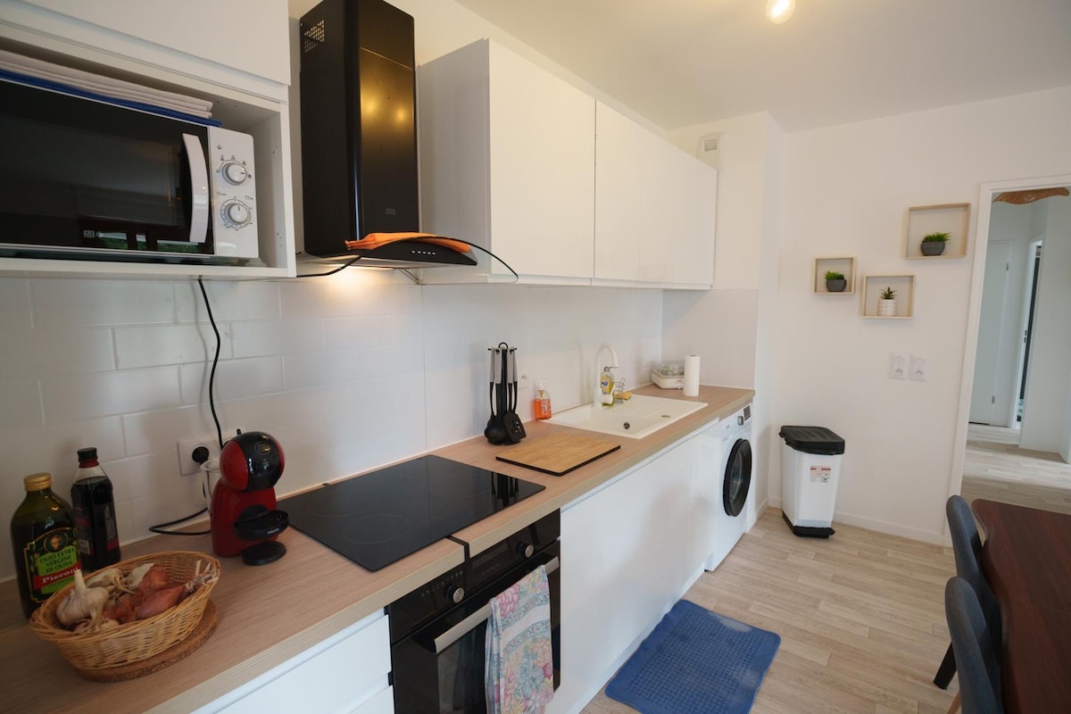 A modern kitchen is presented with sleek white cabinetry and a wooden countertop. Essential appliances are featured, including a microwave, stove, and washing machine. A decorative basket rests on the counter, while small plants are displayed on the wall shelving, adding a touch of greenery.