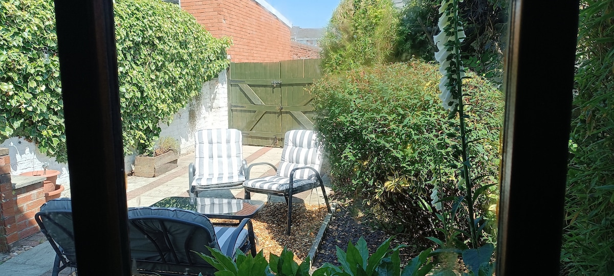 A serene outdoor area features two cushioned lounge chairs and a small table, set against vibrant greenery and a well-maintained garden. A wooden gate leads to a pathway, providing a private and relaxing atmosphere.