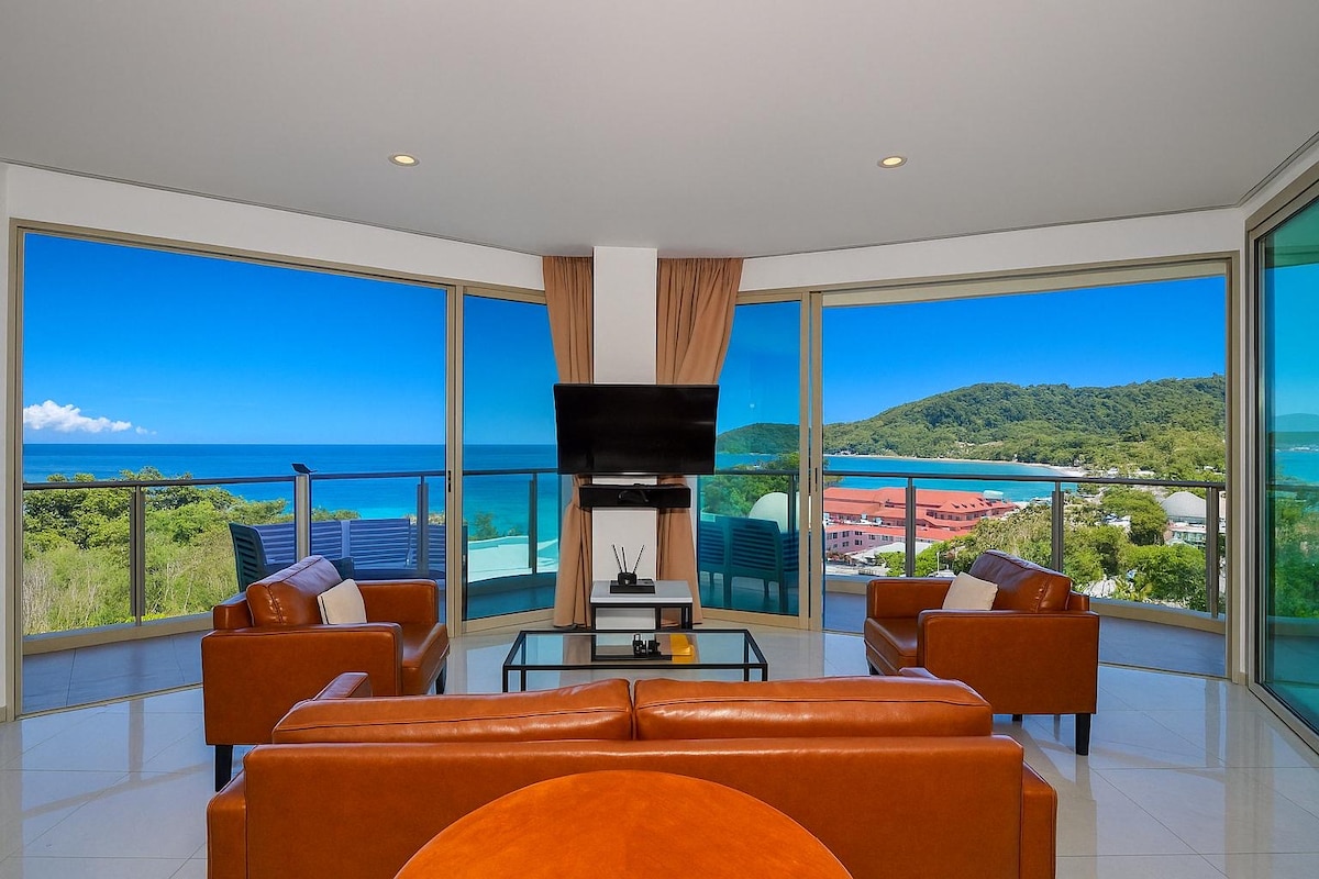A spacious living area is presented with large windows offering wide sea views. Leather seating is arranged around a circular coffee table, and a flat-screen television is mounted on the wall. Natural light fills the room, highlighting the clean, modern design.