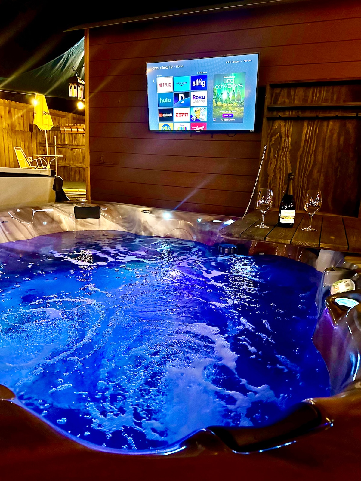 A hot tub with illuminated water creates a relaxing ambiance. A wall-mounted screen displays streaming options for entertainment. Glasses and a bottle of sparkling wine are positioned nearby, suggesting a perfect setting for unwinding. Warm lighting enhances the outdoor space's inviting atmosphere.
