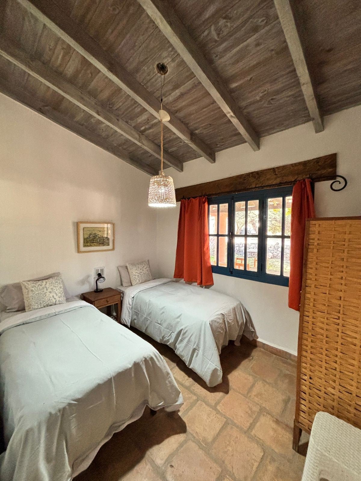 A cozy room features two single beds dressed in light gray bedding, positioned beside a wooden nightstand with a lamp. Natural light filters through a pair of blue-framed windows adorned with orange curtains, highlighting the textured stone floor and wooden ceiling beams.