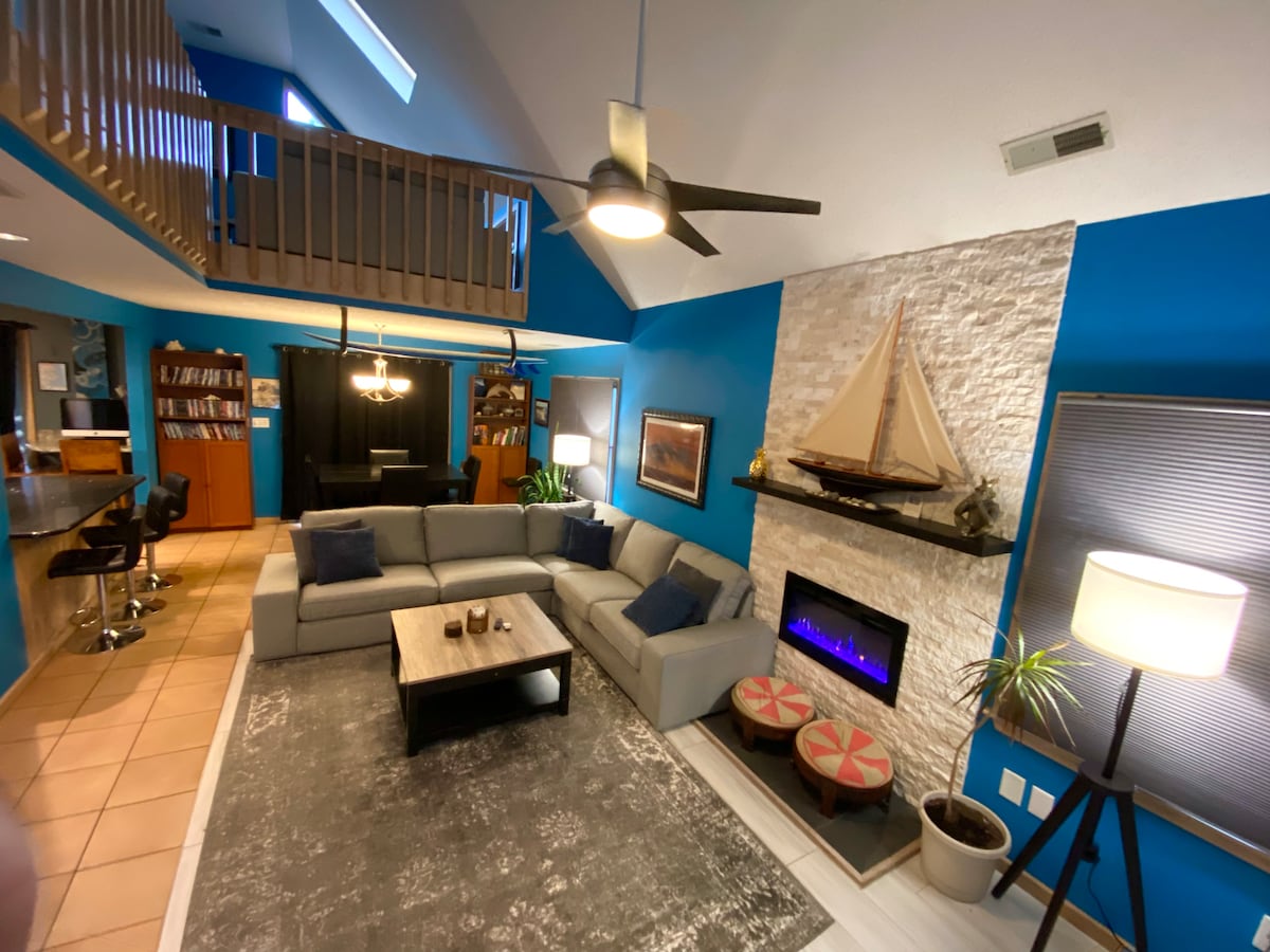 A modern living area features a spacious gray sectional sofa, complemented by dark blue walls. A stylish electric fireplace is set within a stone wall, accompanied by decorative plants and a coffee table. Natural light floods the room through large windows, enhancing the welcoming atmosphere.