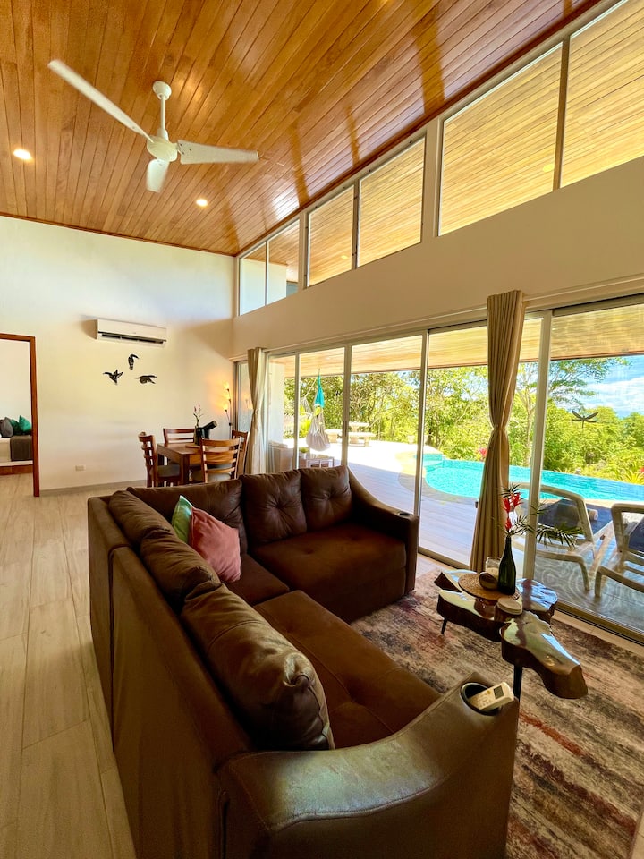 סלון עם חללים פתוחים, דלתות ותריסים נפתחים לבריכה פרטית ולנוף שופע של ג'ונגל. התכונות כוללות טלוויזיה 55 אינץ', ספה מודולרית, ריהוט עץ טיק בעבודת יד, שולחן אוכל וספסלי בר, מושלם למנוחה אחרי יום של סיורים.