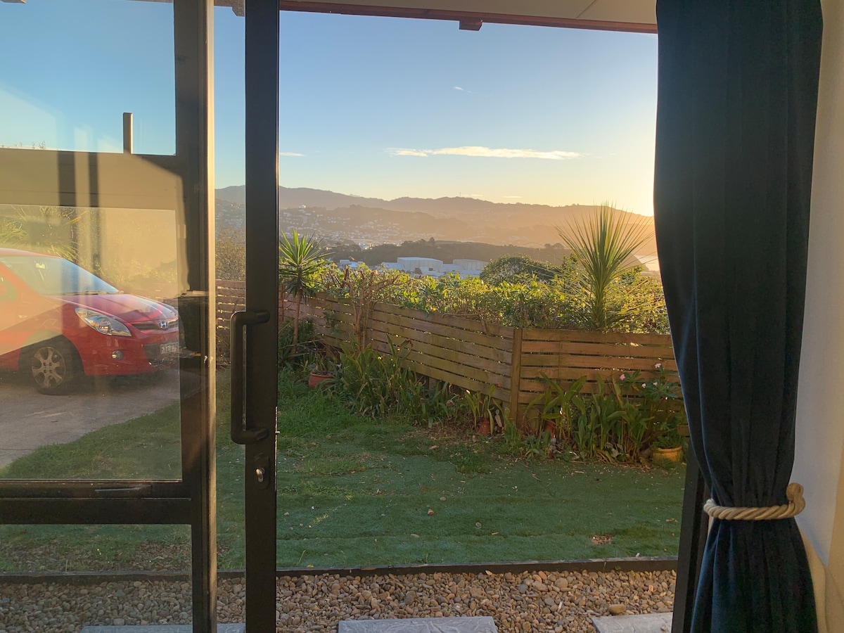 A view from the studio highlights a tranquil outdoor space with greenery and a wooden fence. Sunlight casts a warm glow over the landscape, showcasing distant hills and the valley below. A red vehicle is parked outside, enhancing accessibility to the unit.