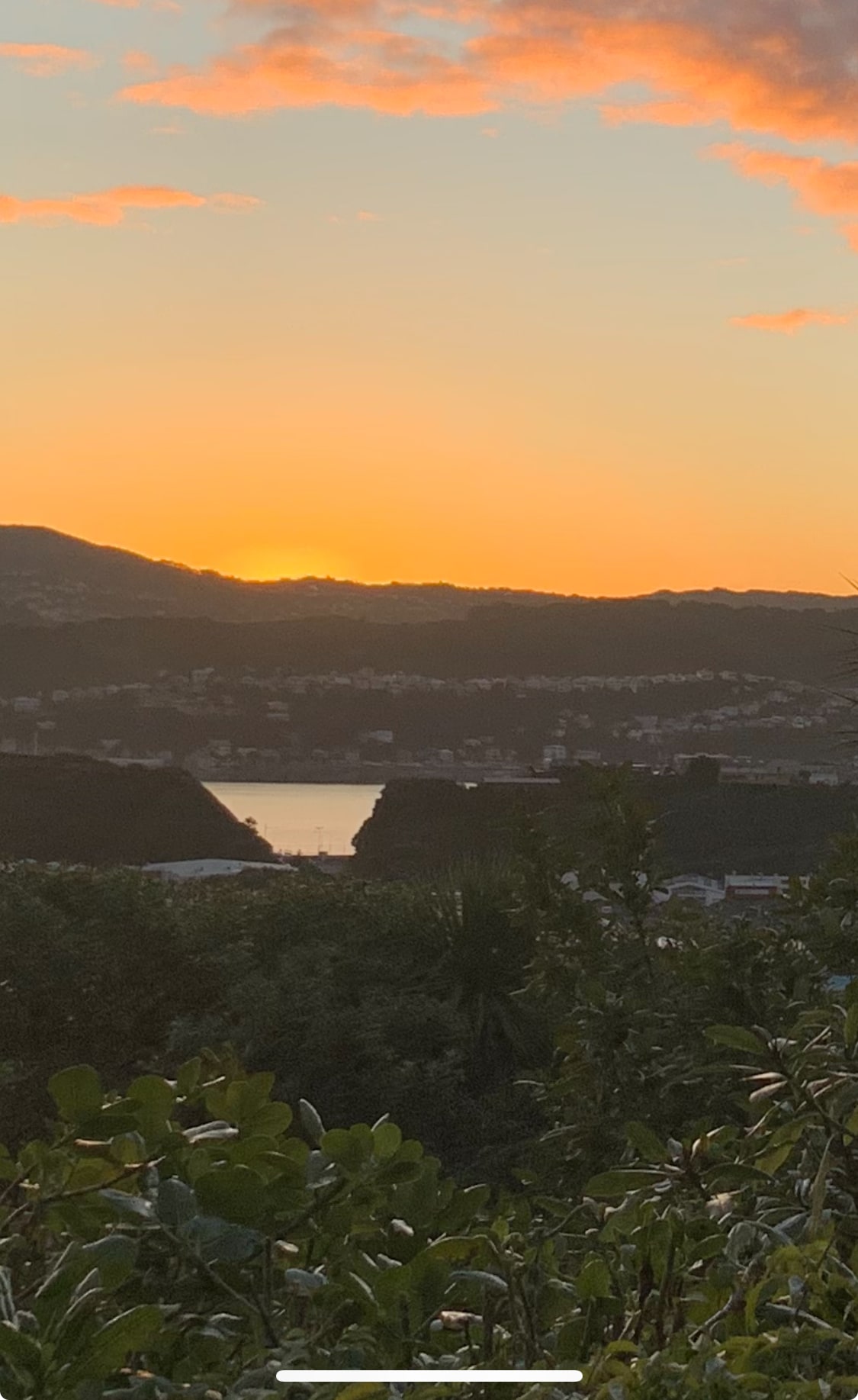 A serene sunset is captured over the landscape, showcasing hills and a body of water in the distance. The warm hues of the sky blend seamlessly with the silhouettes of the surrounding hills, creating a tranquil setting amidst lush greenery in the foreground.