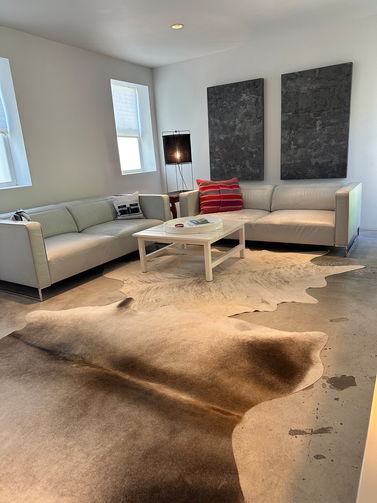 A living area features two light-colored sofas arranged around a central coffee table. A plush area rug adds warmth to the polished concrete floor. Two large artworks adorn the wall behind, while natural light filters through the nearby windows.