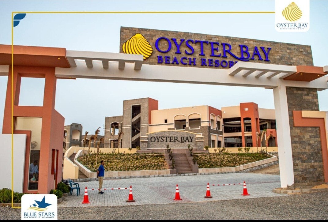 The entrance of Oyster Bay Beach Resort features a large, welcoming sign above a well-paved pathway. Neatly arranged landscaping surrounds the entry, creating an inviting atmosphere. The modern architecture of the resort buildings is visible in the background, highlighting the contemporary design.