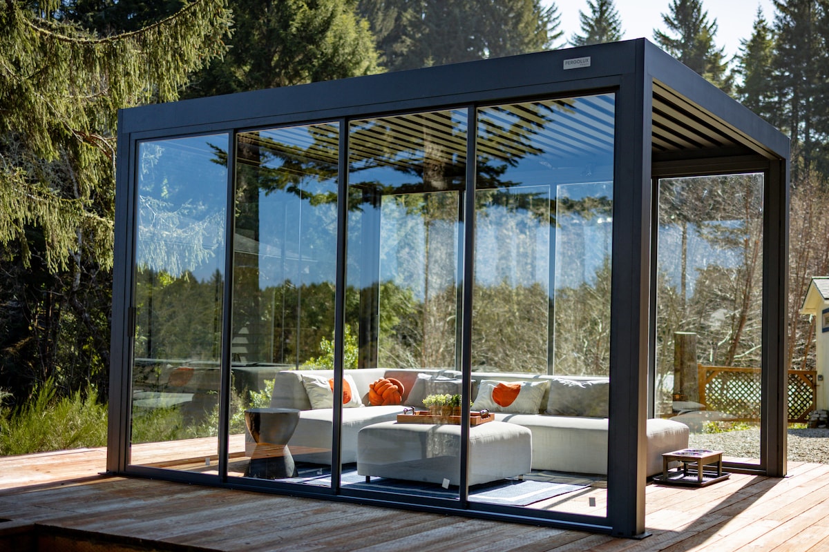 A modern glass enclosure features large transparent walls, providing an expansive view of the surrounding forest. Inside, a comfortable seating area is arranged with a light-colored sofa and orange accent pillows. Natural light floods the space, enhancing its serene atmosphere.