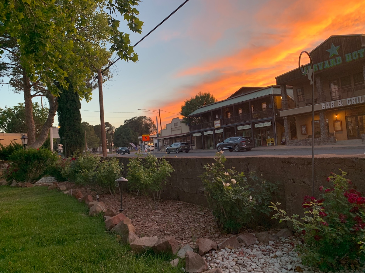 A view of the street showcases a vibrant sunset with warm tones. The foreground features a landscaped area with rocks and shrubs, while the background includes several buildings, including a bar and grill. Streetlights are visible, and cars can be seen parked along the road.