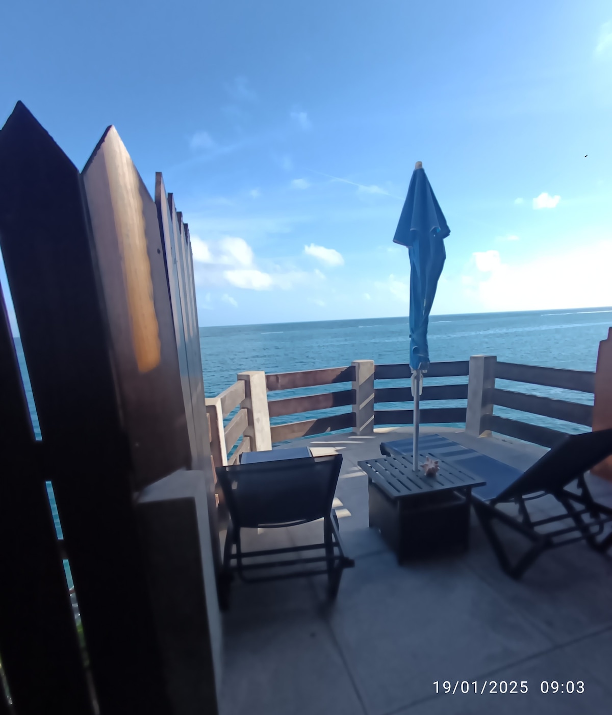 A private terrace extends over the ocean, featuring two lounge chairs and a small table. A blue umbrella provides shade, while the tranquil waters and sky blend seamlessly in the background, creating a peaceful outdoor retreat.