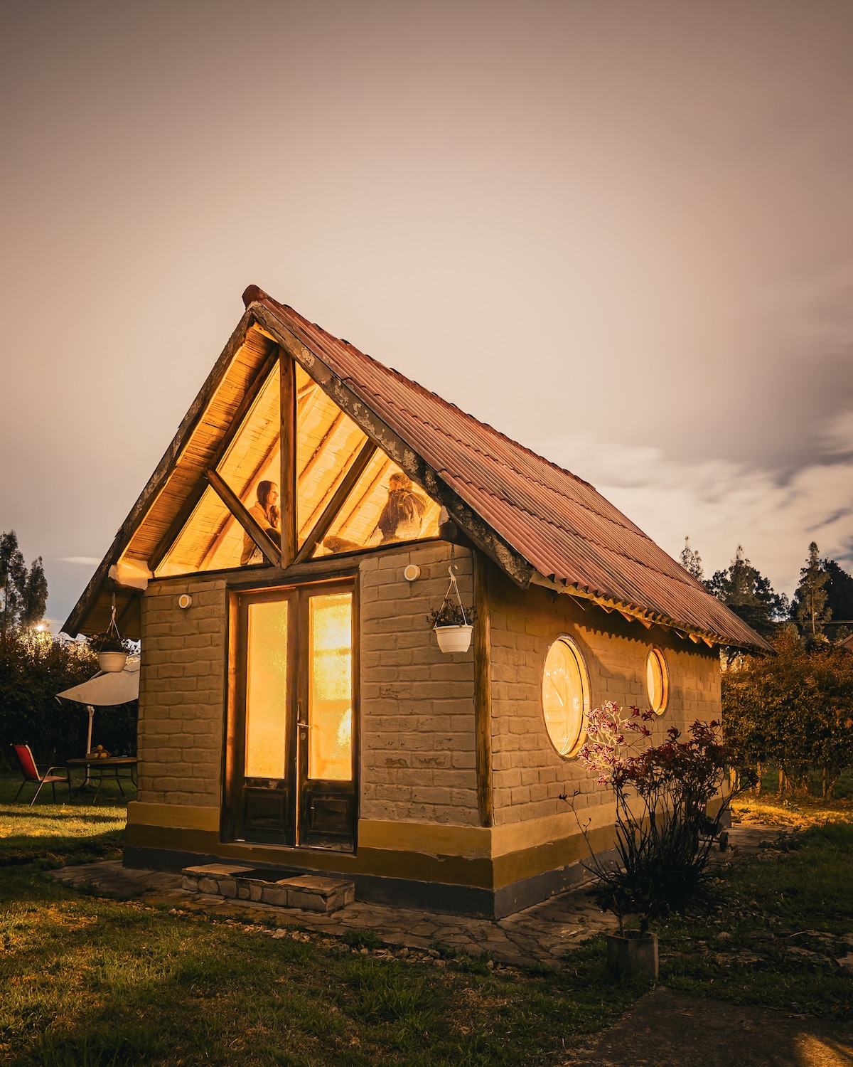 A charming tiny house is highlighted against a nighttime sky, featuring a thatched roof and warm lighting emanating from its windows. The structure showcases rounded windows and a simple stone exterior, with a small garden area in front, contributing to a cozy, inviting atmosphere.