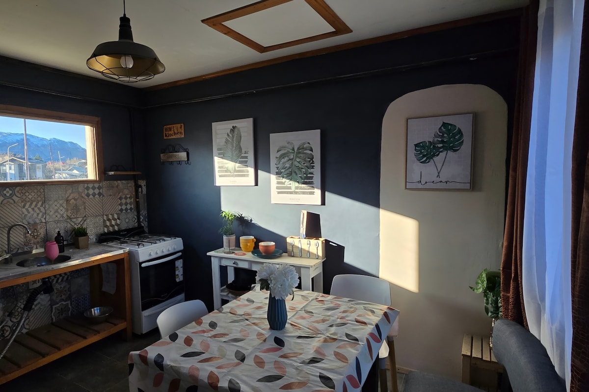 A functional kitchen space is highlighted by a wooden dining table with a floral centerpiece. Soft natural light streams through the window, illuminating wall art featuring leafy designs. A stove and sink are visible along the wall, with a cozy seating area nearby.