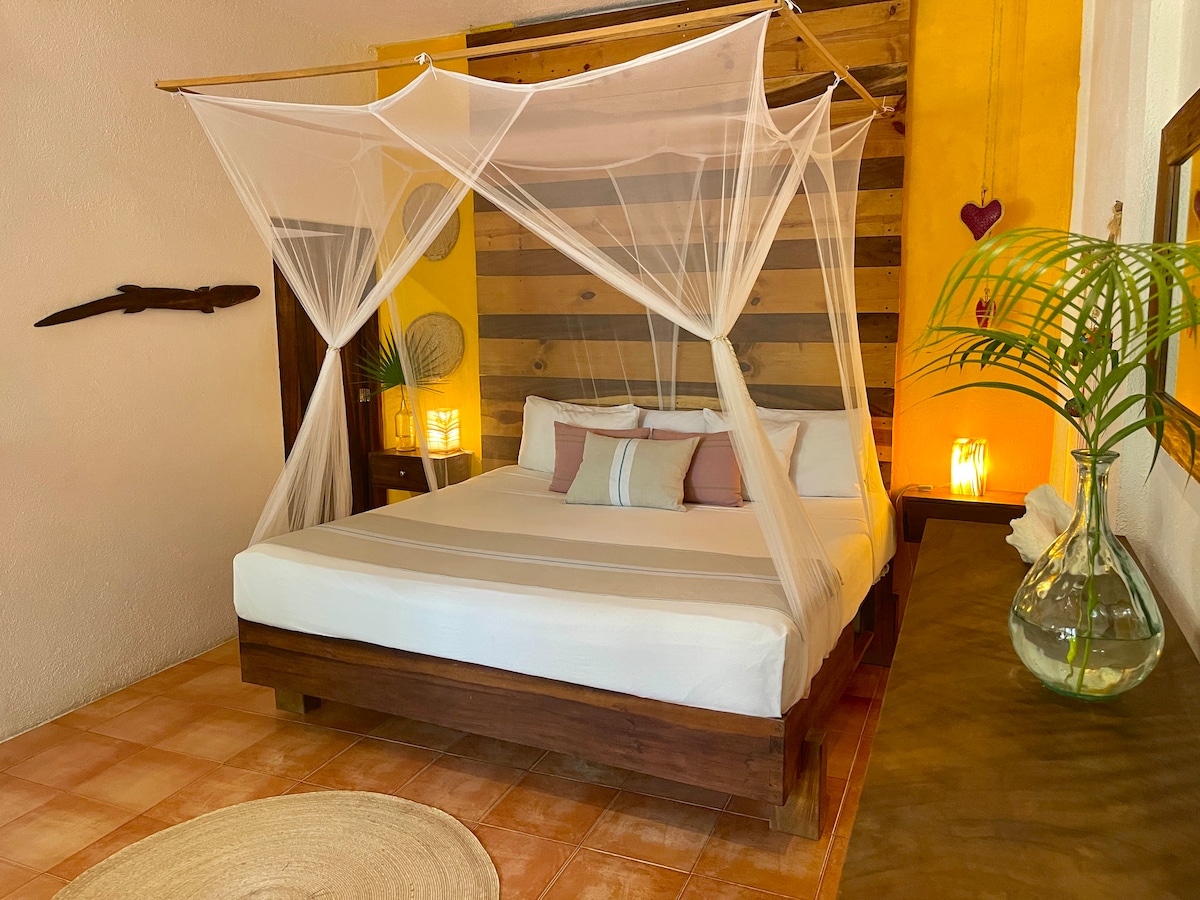 A spacious sleeping area features a king size bed draped with a delicate mosquito net. The warm-colored walls are complemented by natural wooden accents, and soft lighting creates a calming atmosphere. A round rug lies beneath the bed, adding a touch of texture to the floor.