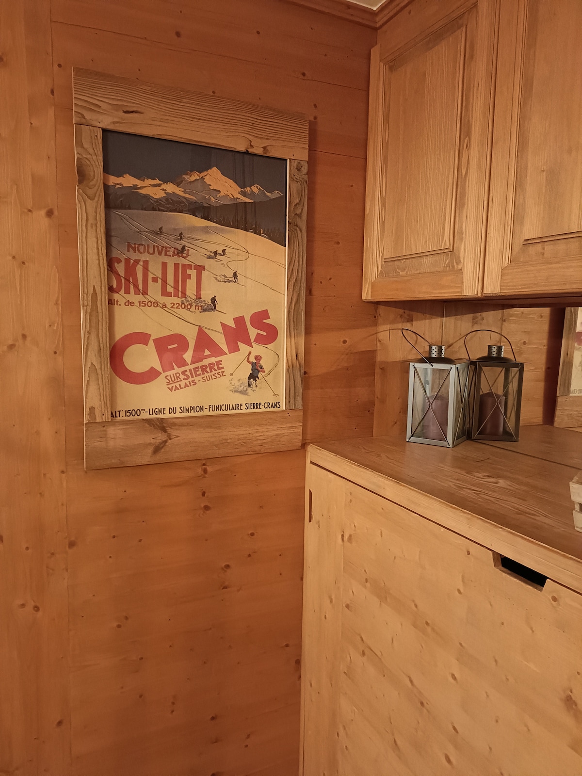 A wooden wall features a vintage ski lift poster promoting Crans, framed in rustic wood. Two lantern-style lights are positioned on a nearby surface, providing subtle illumination. The warm tones of the wood create a cozy atmosphere.