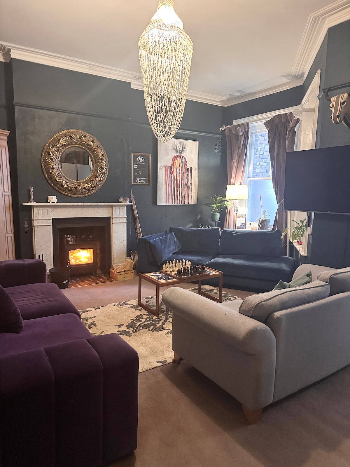 A warm living area features a dark green accent wall, elegant fireplace, and a large mirror. Comfortable seating is provided by a grey sofa and a purple armchair. A chess set adorns the coffee table, with light streaming in from a nearby window.