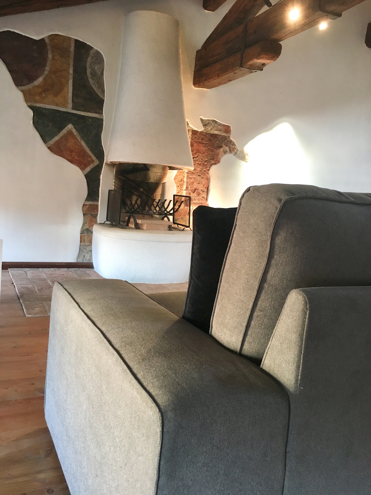 A comfortable sectional sofa is positioned invitingly, with a modern fireplace visible in the background. The fireplace features a unique design, complemented by original frescoes on the wall. Natural light softly illuminates the space, enhancing the atmosphere of the open area.