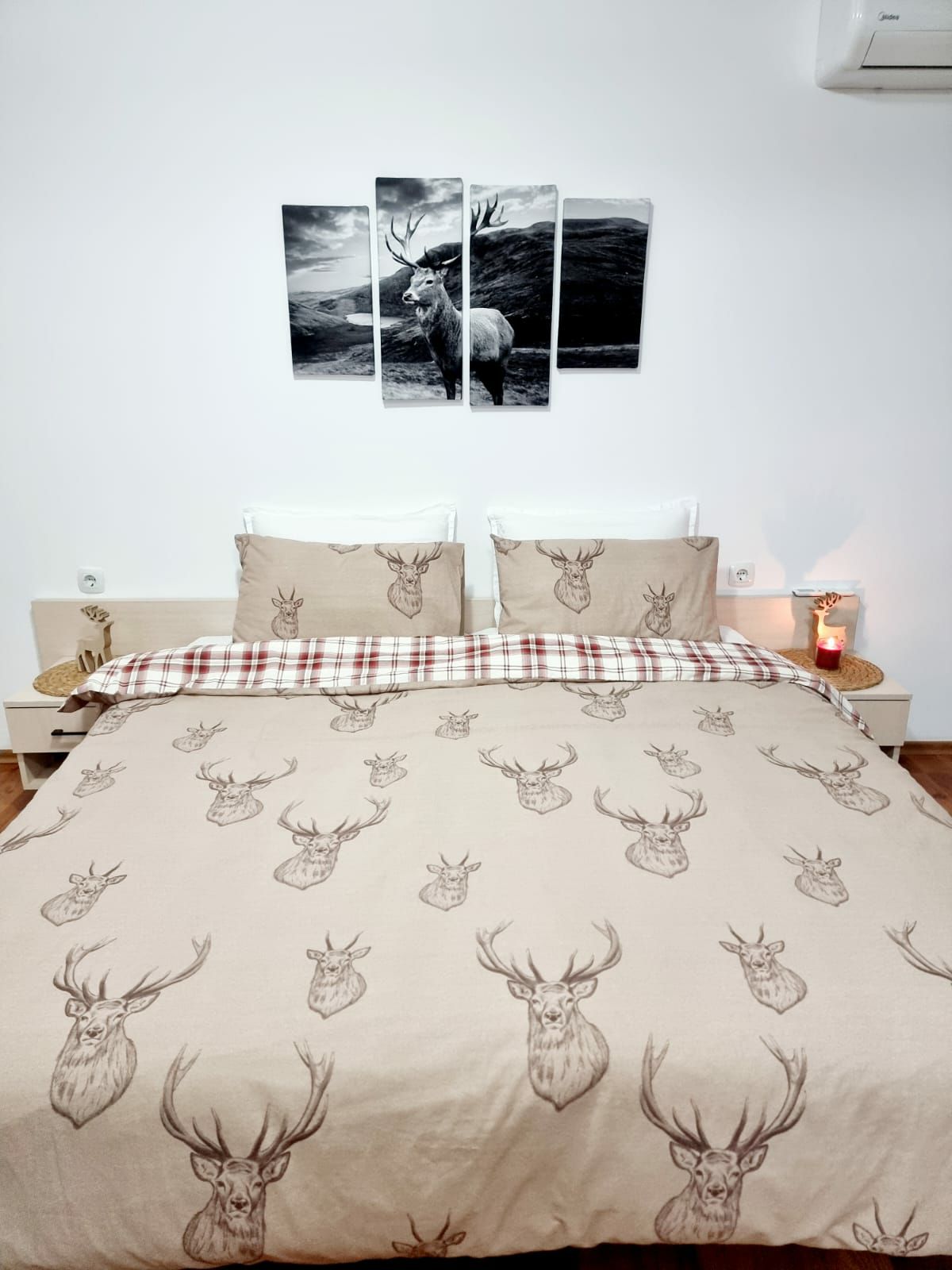 A spacious bed is dressed in a patterned duvet featuring deer motifs. Soft cushions are arranged at the head of the bed, complemented by a plaid throw. A serene photograph of a deer is displayed above the bed, while a candle casts a gentle glow in the corner.