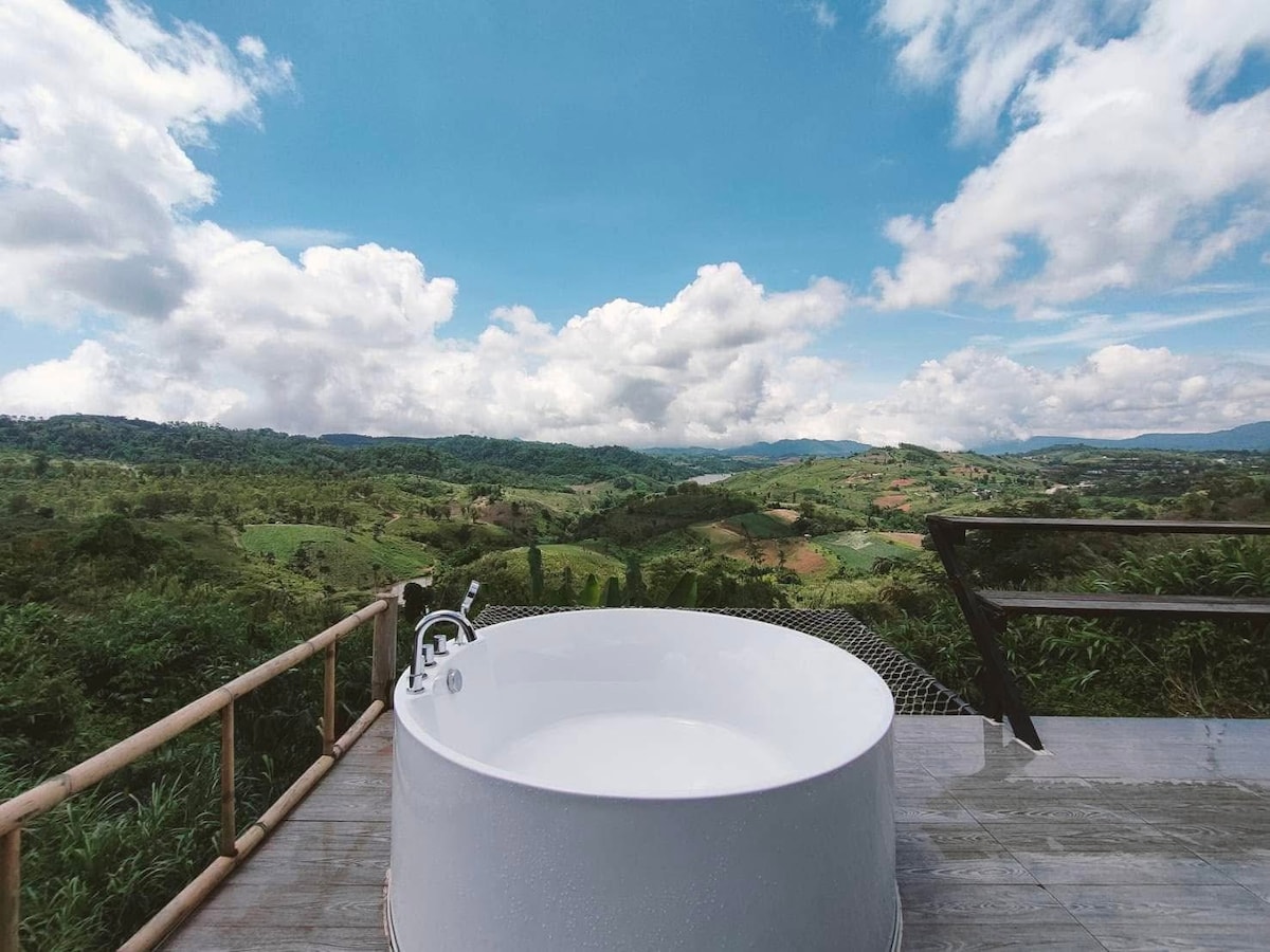 An outdoor hot tub is positioned on a wooden deck, offering views of rolling hills and lush greenery. The sky is partially cloudy, with bright blue spaces accentuating the scenic landscape. Surrounding vegetation adds to the natural ambiance of the setting.