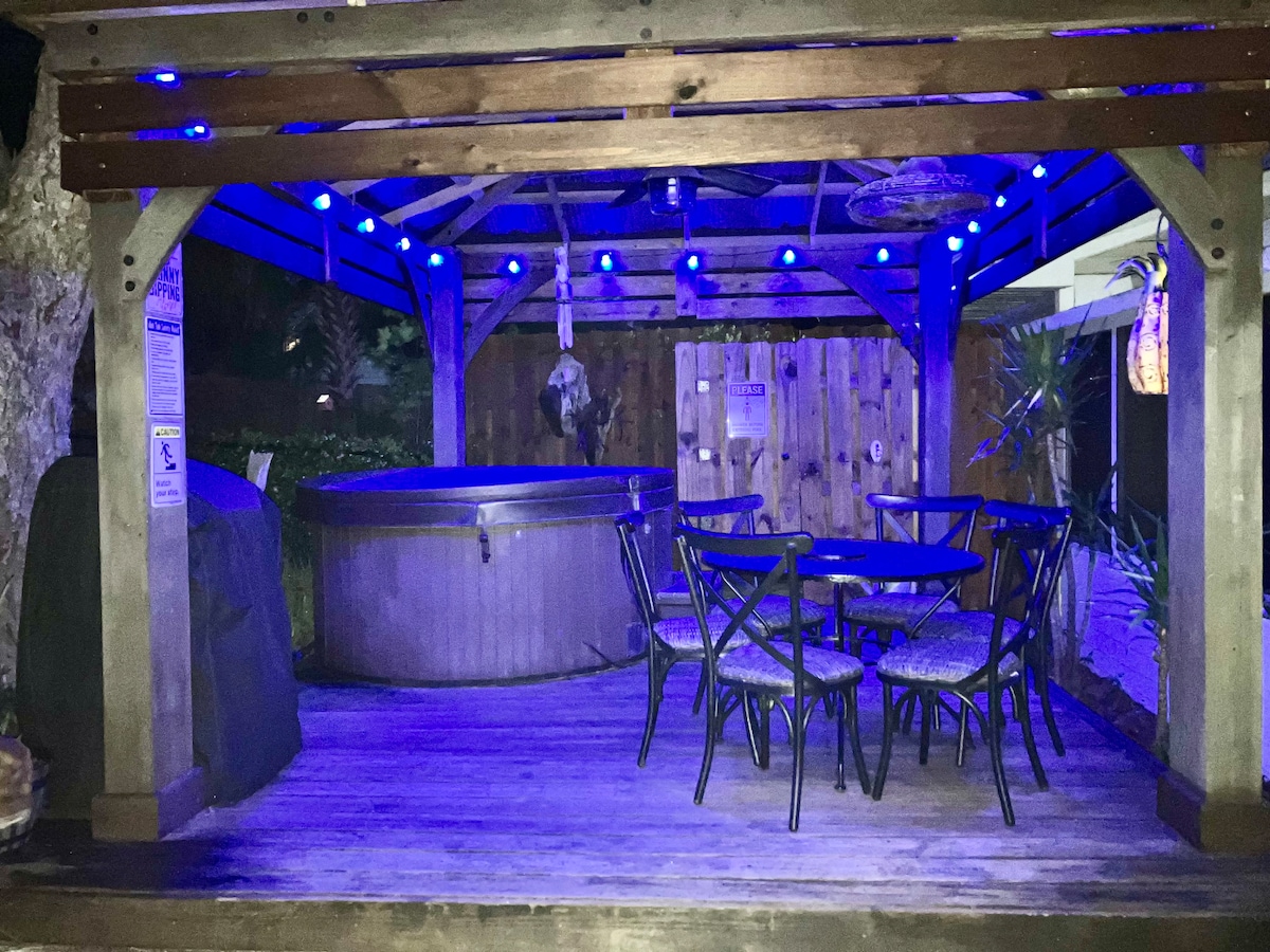 An inviting outdoor space is encompassed by a wooden structure with blue lighting highlighting the area. A spacious hot tub is positioned to one side, while a round table with four chairs is available for seating. A wooden privacy fence creates a secluded atmosphere.