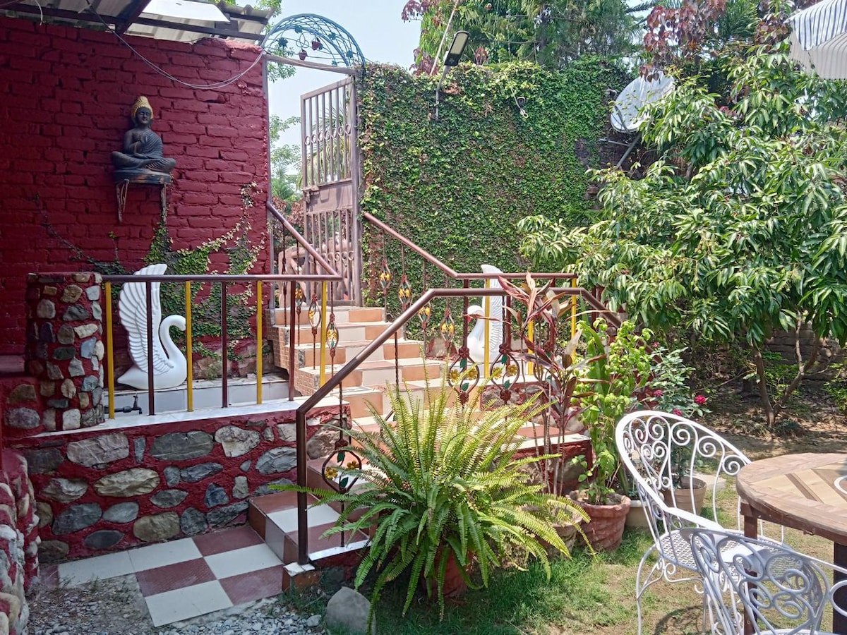 A charming entrance is surrounded by lush greenery and colorful plants. Stairs lead to an upper level, flanked by a decorative iron railing. A stone pathway adds texture, while a small seating area features a white wrought-iron chair, inviting relaxation in this tranquil garden setting.