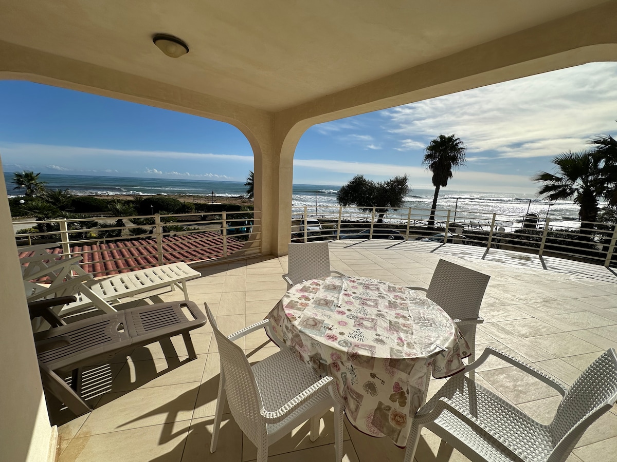 An expansive veranda offers a scenic view of the beach and sea. A round dining table is set with a floral tablecloth, surrounded by white chairs. Sunlight reflects off the water, while palm trees are visible in the background.