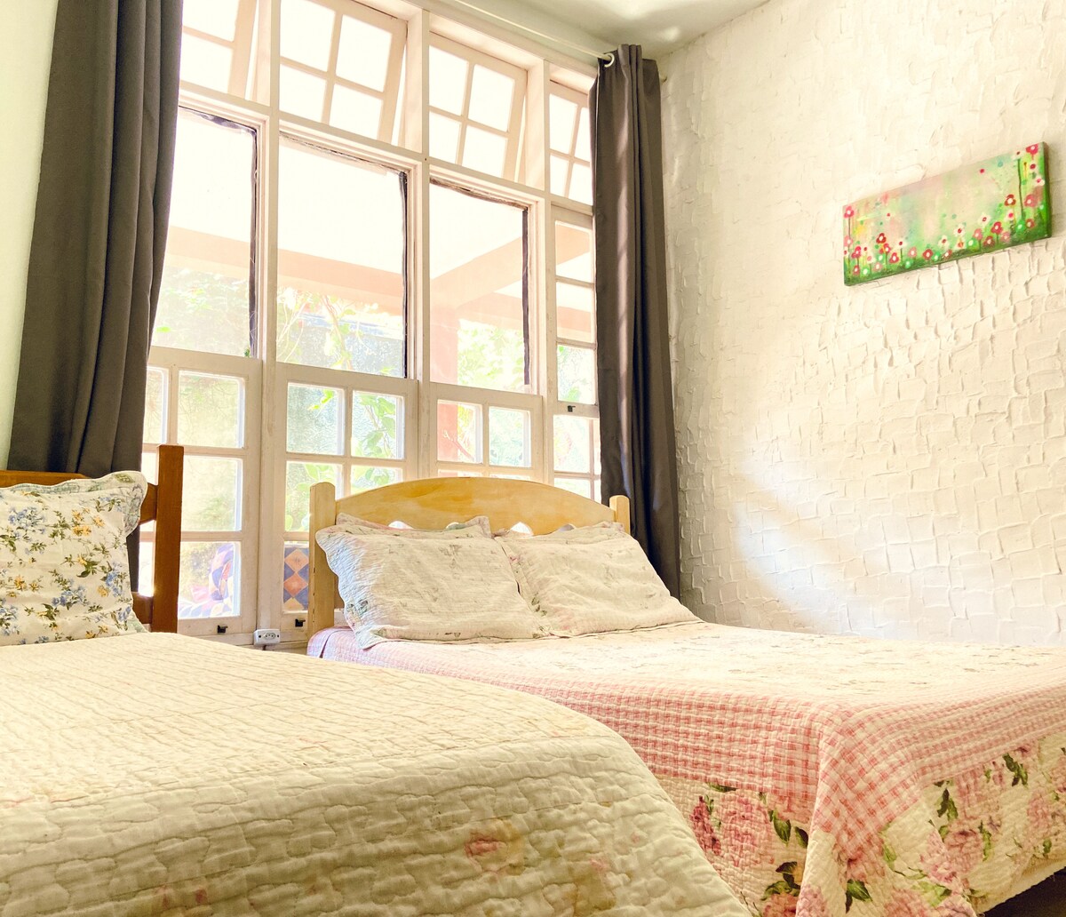 A spacious bedroom is featured, containing a double bed and a single bed. Large windows provide natural light, with soft curtains framing the view. The walls are textured, enhancing the room's character, and a decorative artwork hangs above the beds, adding a touch of color.