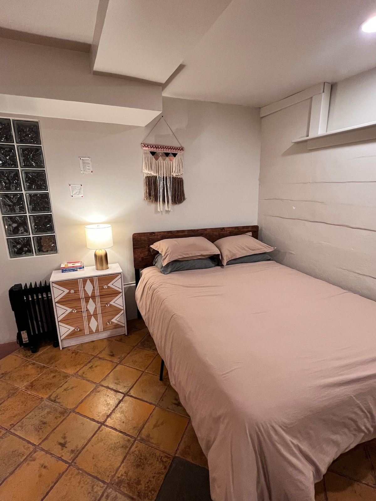 A cozy sleeping area is presented with a spacious bed covered in a soft beige comforter. A decorative wall hanging adds visual interest above the bed, while a small bedside table with a lamp provides soft illumination. Textured tile flooring enhances the warm, inviting atmosphere.