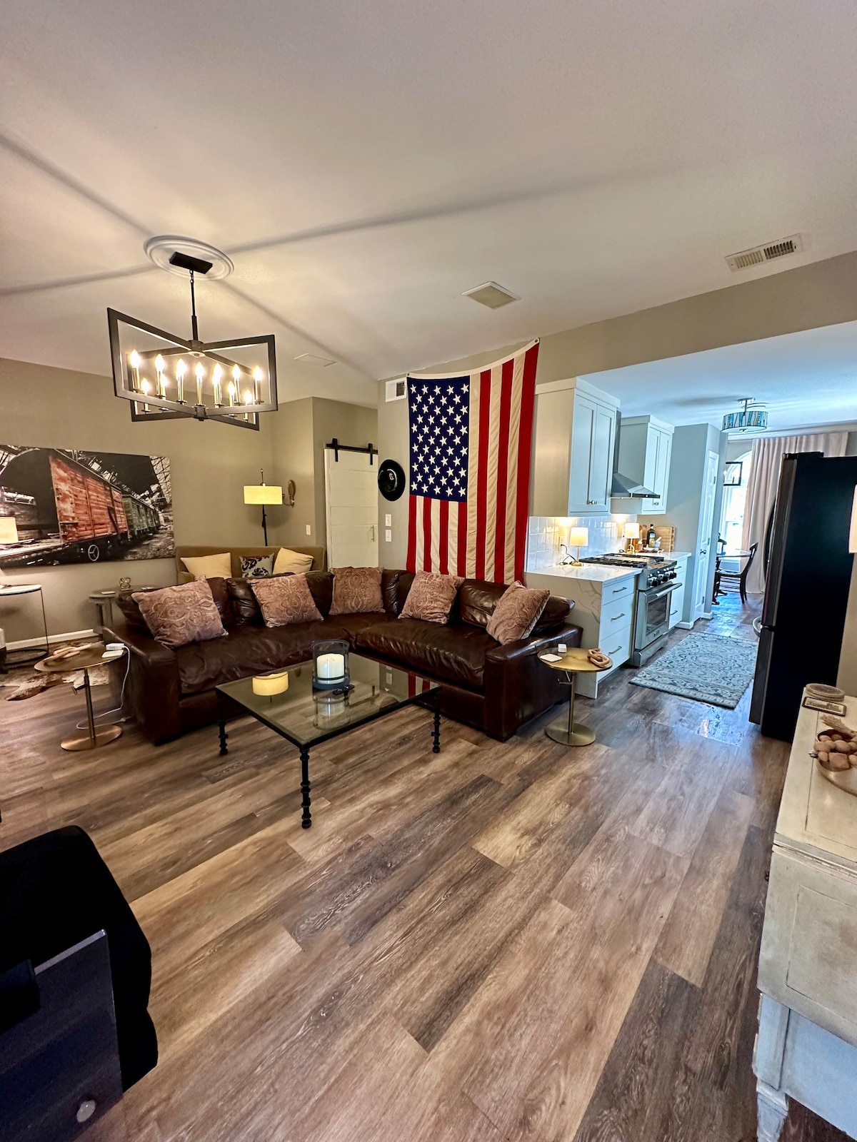 A cozy living area is highlighted by a brown sectional sofa adorned with decorative pillows. An American flag is displayed on the wall behind the sofa. The adjoining kitchen is visible, featuring modern appliances and ample lighting, creating a welcoming ambiance.