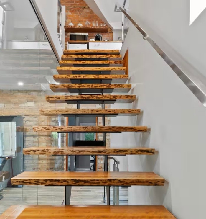 A modern glass stairwell showcases floating wooden steps with natural grain patterns, leading to an upper level. Exposed brick walls are visible in the background, complementing the contemporary design. Good lighting highlights the textures of the materials used.