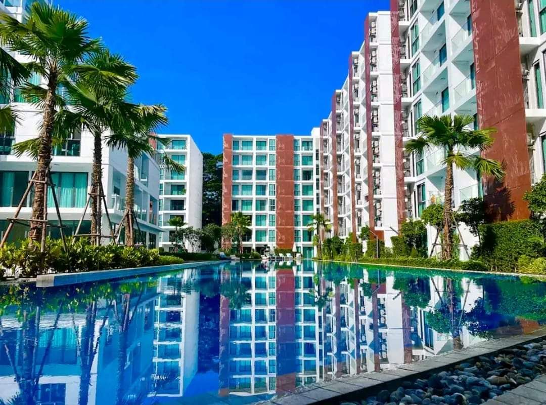 A large, reflective swimming pool is framed by vibrant greenery and palm trees, with modern multi-story buildings visible in the background under a clear blue sky.
