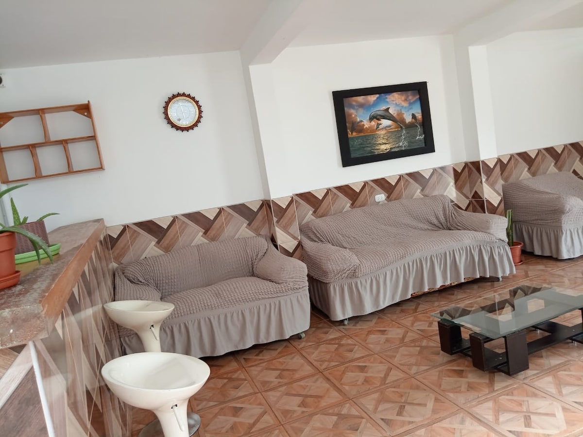 The lounge area is furnished with two upholstered sofas with light-colored covers, complemented by a glass coffee table. A wall clock and framed artwork depicting dolphins are displayed above a neatly tiled surface, enhancing the inviting and relaxed ambiance.