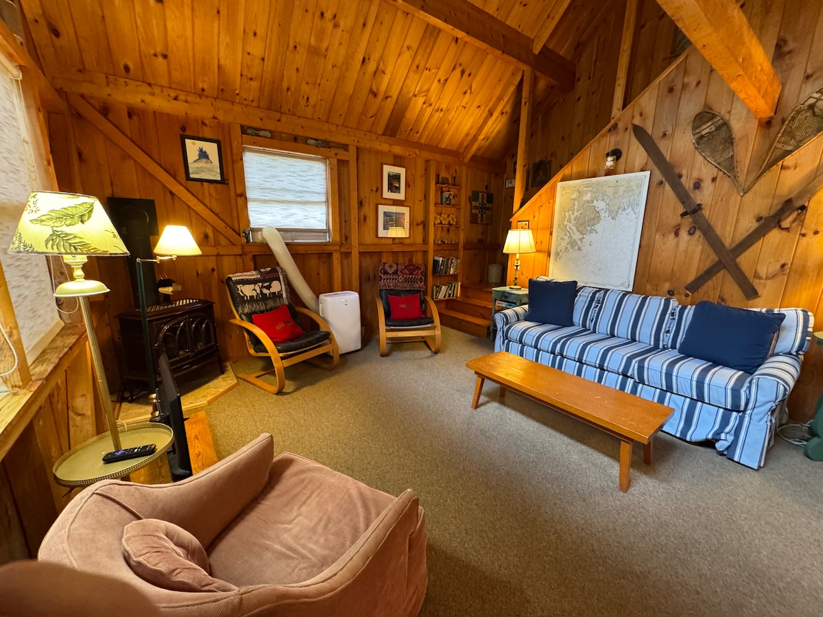 The cabin's living area features a wooden interior with high ceilings. A striped sofa is positioned across from a cozy chair and a small coffee table. Two lamps provide soft lighting, while wall decor, including a map and decorative oars, enhances the rustic charm.