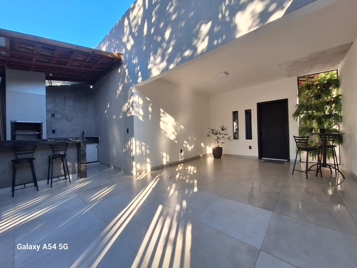 An open and airy outdoor space features smooth tiled flooring and striking shadows cast by overhead foliage. A small dining table with chairs is positioned near the entrance, while a barbecue area is visible in the background against a textured wall.