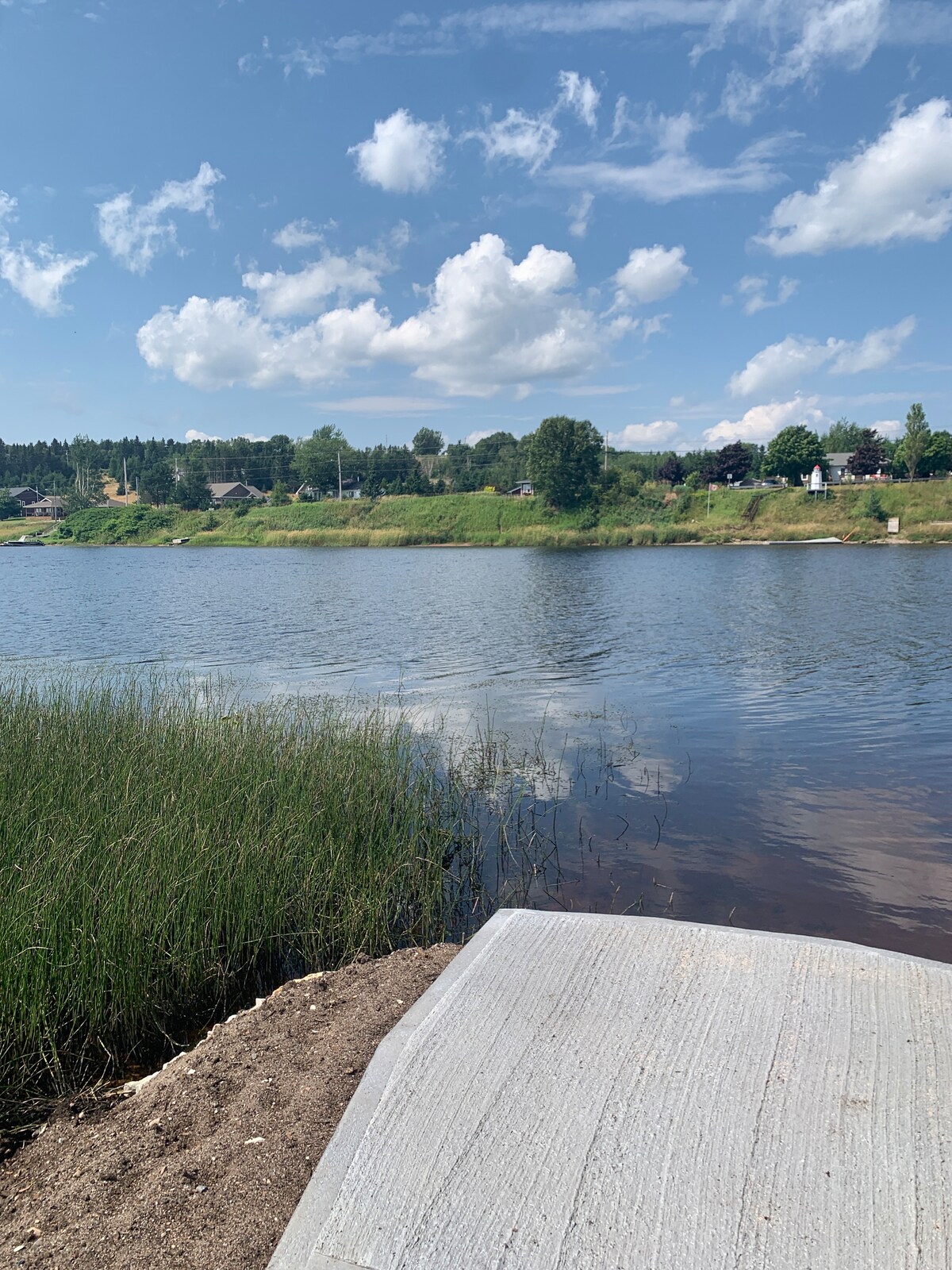 The image captures a tranquil view of the Humber River, with lush greenery lining the banks. Sunlight reflects off the water's surface, while fluffy clouds drift in a blue sky. A concrete platform extends towards the water, framed by tall grass and sandy areas.