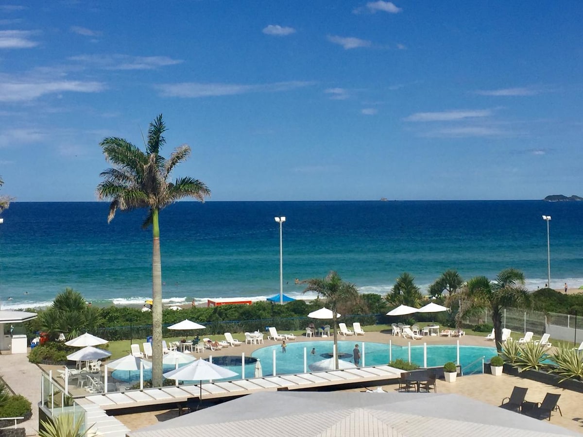 A clear view of the ocean complements a serene outdoor pool area. Loungers and umbrellas are positioned around the pool, with palm trees enhancing the coastal atmosphere. The expanse of blue water stretches to the horizon, inviting relaxation and leisure.
