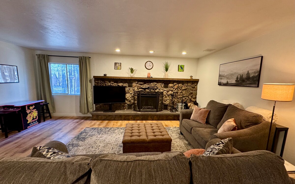 A welcoming living area features a stone fireplace centered on a wooden wall. A comfortable seating arrangement includes a sofa and chairs, with soft throw pillows. A quilted ottoman sits in the middle of the room, complemented by warm lighting and a large window showcasing forest views.