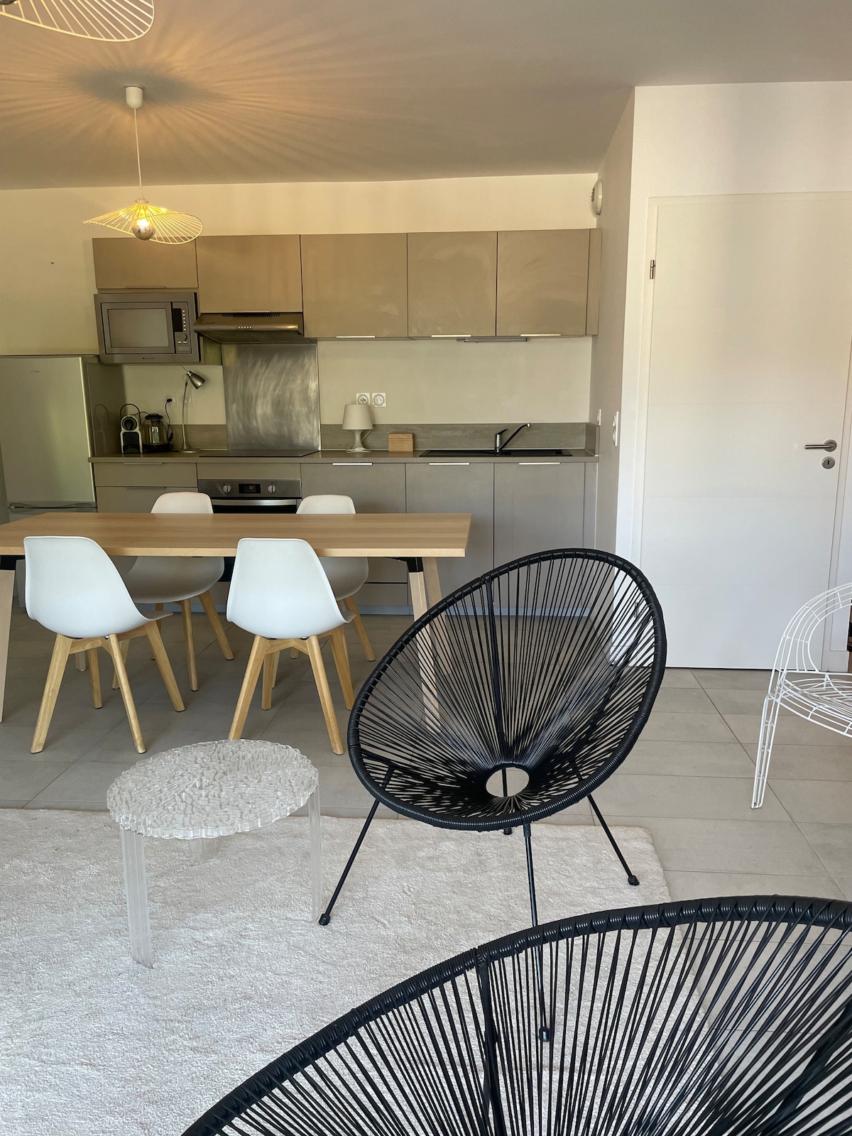 A modern kitchen and dining area are visible, with a wooden table surrounded by stylish chairs. A sleek, stainless steel refrigerator and appliances complement the contemporary design. Natural light fills the space, enhancing the neutral color palette throughout the apartment.