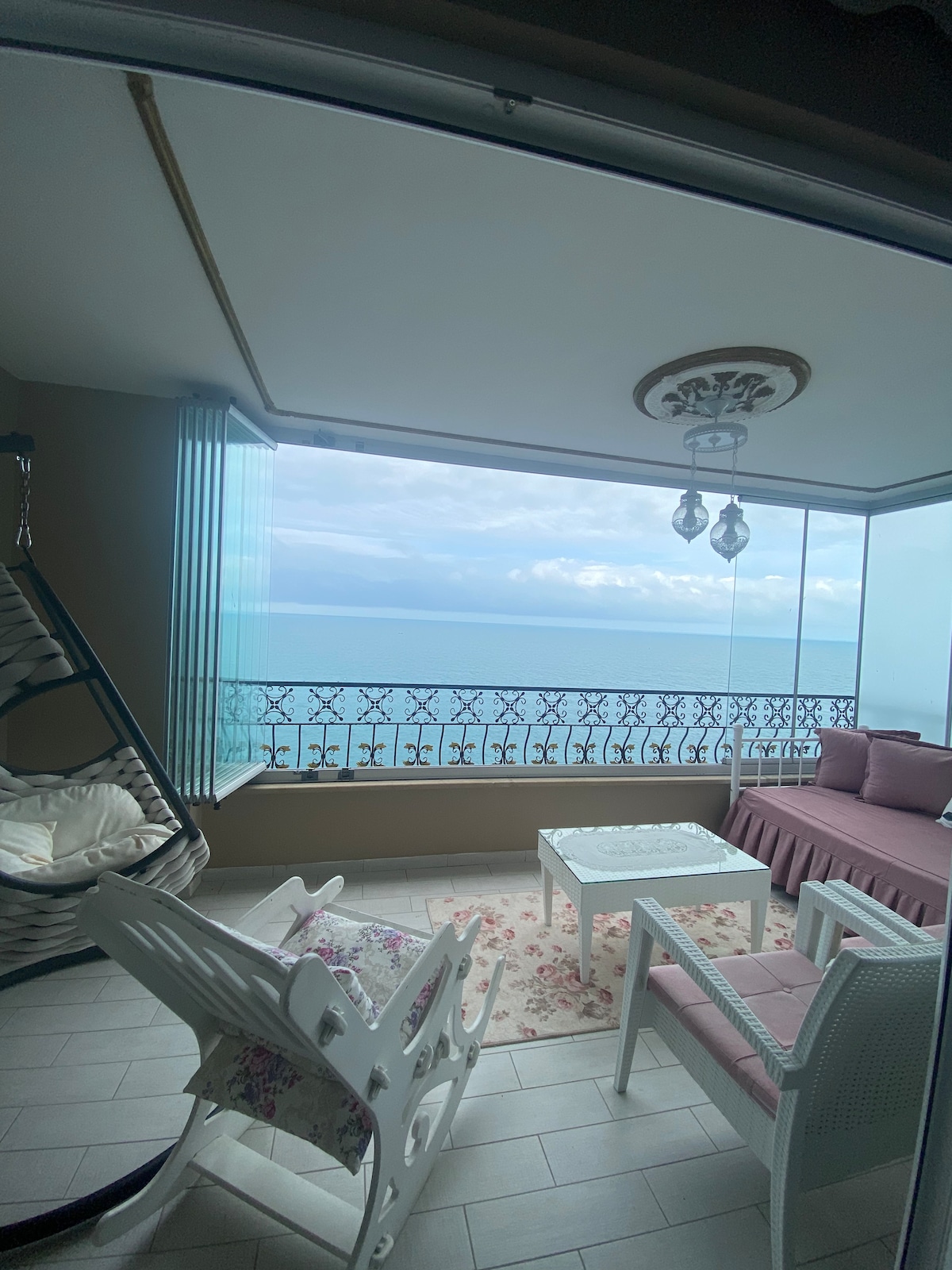 A bright and airy space features a cozy seating area with a sofa and two comfortable chairs. A decorative table is positioned in the center, while large windows offer an unobstructed view of the sea, enhancing the serene ambiance.