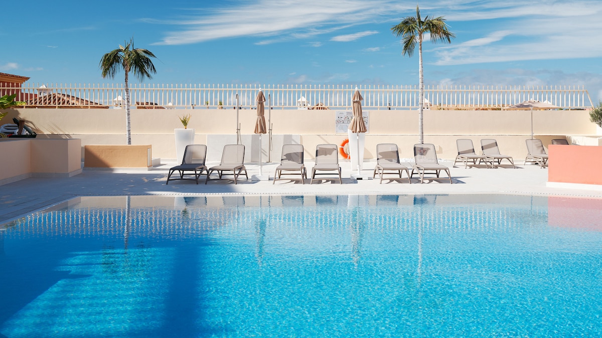 A clear blue swimming pool is surrounded by several lounge chairs under tall palm trees. Umbrellas are positioned for shade alongside the pool, and a decorative planter adds a touch of greenery to the scene. The sky is bright with few clouds.