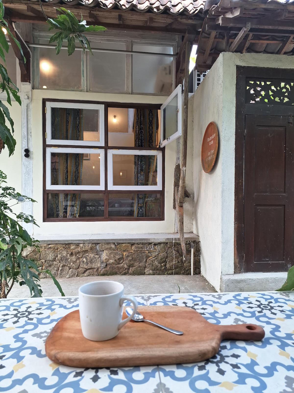 A wooden cutting board is set on a patterned table, featuring a white mug and a spoon. In the background, a traditional wooden structure with multiple windows is visible, showcasing colorful curtains and inviting warm light.