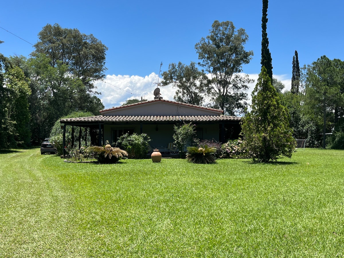 A charming countryside house with a traditional style is set amidst lush greenery. A wide porch adorned with flowering plants enhances the outdoor appeal. The landscape features a manicured lawn leading to the entrance, framed by trees that provide a sense of privacy.