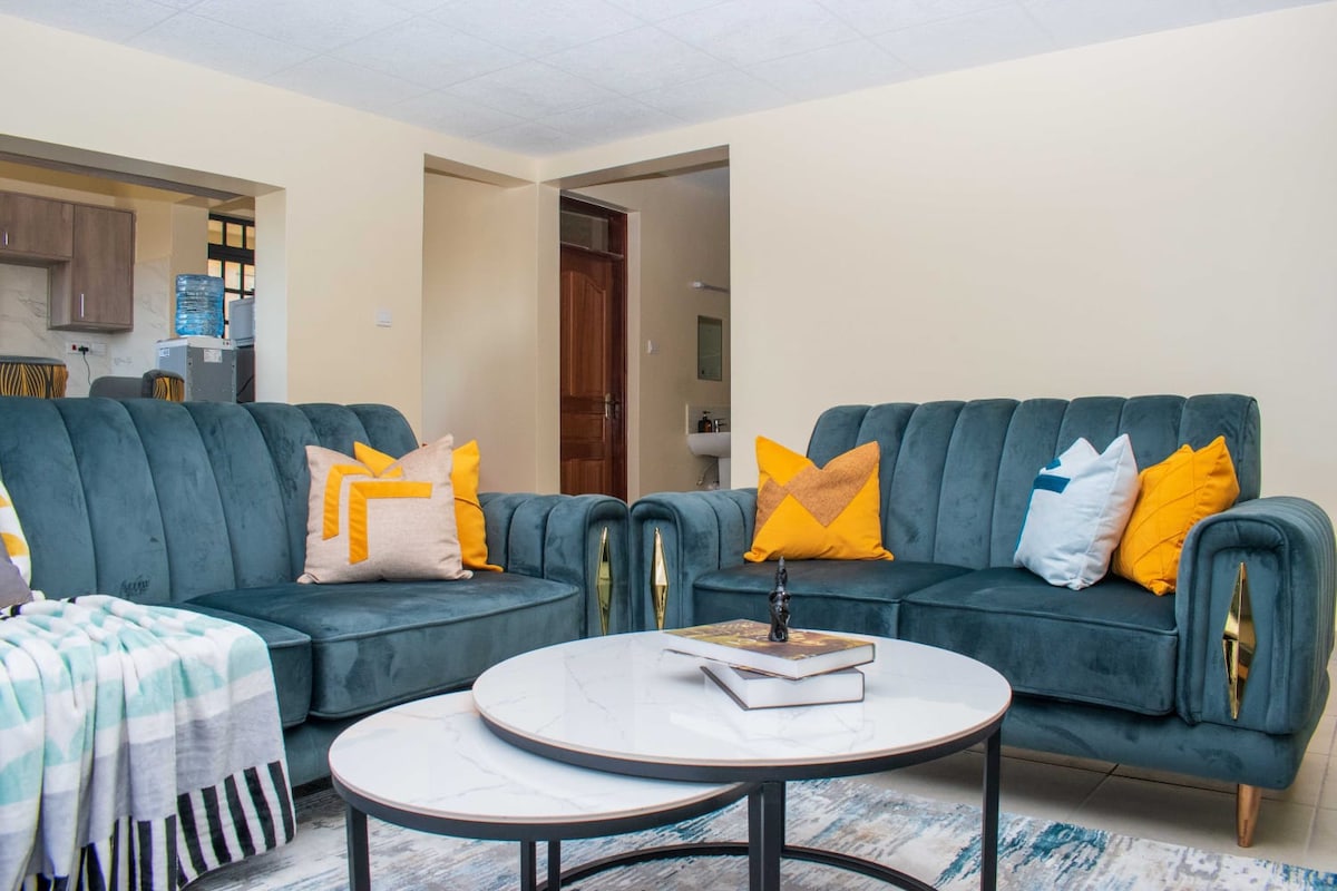 Two plush sofas in soft teal are positioned around a round coffee table. The sofas are adorned with decorative pillows in shades of yellow and white. A light-toned rug is laid beneath, while a warm neutral wall complements the cozy seating area.